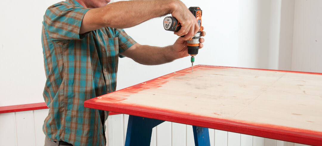 Die Holzplatte mit drei Schrauben auf jeder Seite auf dem Gestell der Beine fest schrauben, für eine bessere Stabilität und Haltbarkeit des Tisches beim Toben und Spielen.